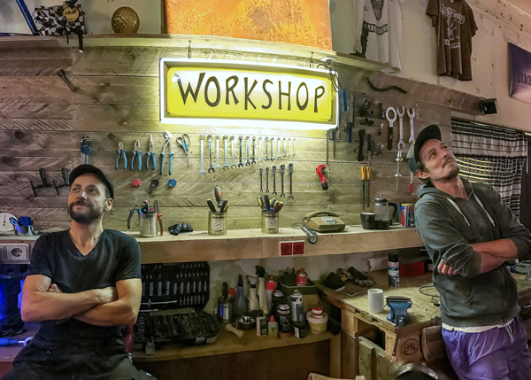 Zwei Männer stehen lächelnd und mit verschränkten Armen in einer Fahrradwerkstatt. Im Hintergrund Werkzeuge und eine Leuchtbox, auf der „Workshop“ steht.